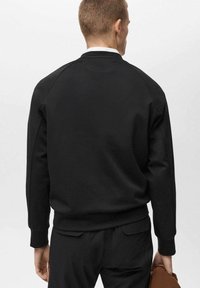 Felpa nera con maniche raglan e polsini a coste. Texture liscia, design minimale. Indossata con pantaloni scuri e in mano una borsa marrone.