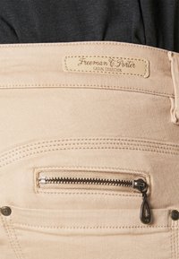 Jean beige avec une poche zippée discrète, étiquette en cuir beige portant l'inscription "Freeman T. Porter Casual Collection", et détails de couture subtils.