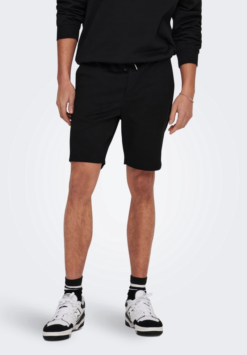 Persona che indossa pantaloni corti neri, una maglietta a maniche lunghe nera, sneaker bianche e nere e calze nere, in piedi contro uno sfondo semplice.