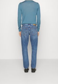 Jeans de mezclilla azul con corte recto, que cuentan con dos bolsillos traseros y un parche de cuero marrón en la cinturilla, combinados con una blusa de manga larga en color azul verdoso.