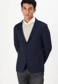 Navy blazer with two buttons, side pockets, and a textured fabric layered over a light beige sweater. Model stands against a white background.