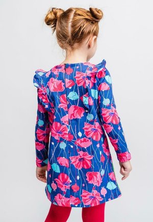 Ung pige med rødt hår i to knuder iført en blå kjole med pink blomster mønster og røde strømpebukser, vendende væk mod en ensfarvet baggrund.