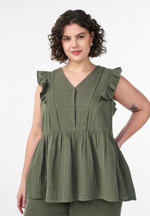 Žena s kučeravými vlasmi nosí bezrukávovú olivovo zelenú blúzku s volánmi a gombíkmi, v kombinácii so zladenými nohavicami, pózuje s rukou na páse.