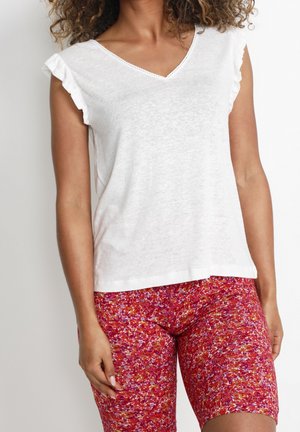 Kobieta ubrana w biały top z marszczonymi rękawami V-neck i dopasowane czerwone szorty w kwiatowy wzór, stojąca na jednolitym tle.