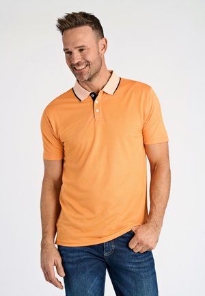 Camisa polo naranja con un cuello crema en contraste y ribetes negros, con mangas cortas y una tapeta de tres botones. Llevada con jeans azules.