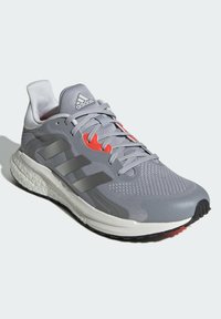 Šedá sportovní bota s prodyšným svrškem, syntetickými detaily, červeným logem a polstrovanou bílou podrážkou. Obsahuje šněrovací uzávěr a texturovaný povrch.