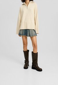 Beiger Strickpullover mit einem Halbres öffner-Kragen, kombiniert mit einem Jeans-Minirock. Braune knielange Stiefel mit Schnallenakzenten und einem runden Zehendesign.