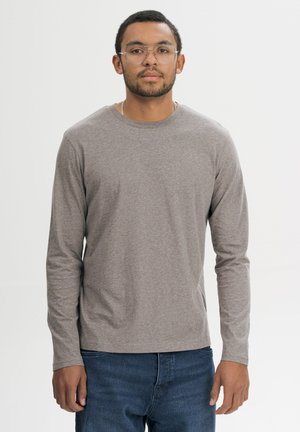 Man draagt een bril met heldere glazen, een grijs shirt met lange mouwen en een blauwe spijkerbroek, staand tegen een effen witte achtergrond.