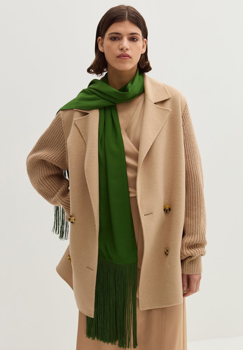 Beige Außenbekleidung mit einem strukturierten Design und gerippten Ärmeln, kombiniert mit einem langen grünen Schal mit Fransen. Glatte Stofftextur.