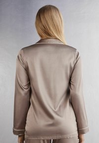 Satinpaja-top i ljus taupe med långa ärmar, en notchad krage och subtil vit piping längs kragen och ärmsluten. Slät textur.