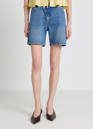 Donna che indossa pantaloncini di jeans blu di media lunghezza, una maglietta gialla e tacchi scuri con punta appuntita, in piedi davanti a uno sfondo semplice.