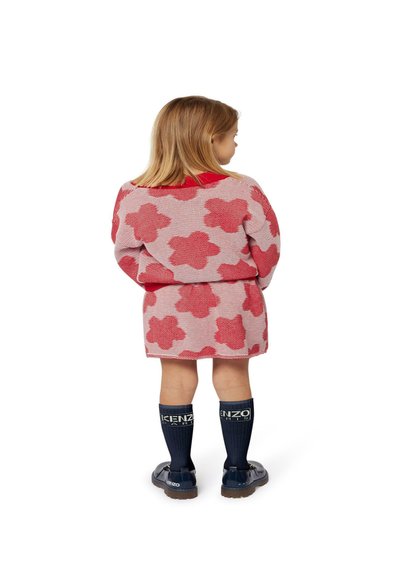 Robe tricotée à motif floral rouge avec des manches courtes, associée à des chaussettes soudées navy jusqu'au genou avec le logo de la marque et des chaussures noires brillantes.