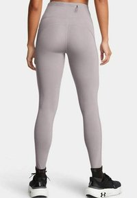Vue arrière d'une personne portant des leggings taille haute mauves et des chaussures de sport noires, sur un fond clair et uni.