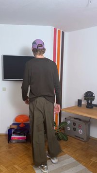 Tmavý top s dlhým rukávom a olivovo zelené široké nohavice. Izba má televízor, oranžovú lampu a drevený písací stôl s dekoráciami a rastlinou.