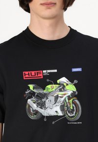 Zwarte t-shirt met een grafiek van een groen-witte motorfiets, voorzien van details en rode tekst met het merk "HUF CBD1000RR."