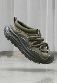 Αθλητικό παπούτσι ανοιχτού ελαιόχρωμου με παχύ, υφασμένο πέλμα και ελαστικά κορδόνια σε βρόχο, αιωρούμενο πάνω από φωτεινό μαρμάρινο πάτωμα και λευκό τοίχο με πάνελ.