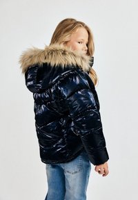 Nafučana tamnoplava jakna s kapuljačom obloženom krznom, sa sjajnom površinom, rebrastim manžetama i kviltanim uzorkom, kombinirana s svijetloplavim trapericama.