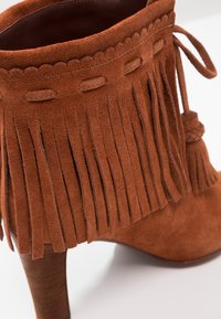 Brązowy zamszowy botek za kostkę z frędzlami, falbaniastymi krawędziami i wysokim obcasem. Posiada plecioną wiązankę w okolicy kostki oraz teksturowaną powierzchnię.