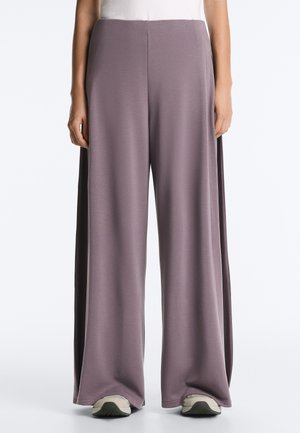 Persona che indossa pantaloni larghi color malva con pannelli laterali neri e scarpe da ginnastica bianche, in piedi davanti a uno sfondo neutro.