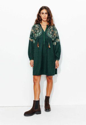 Robe verte brodée avec des manches longues, décolleté en V et des franges à l'avant. Associée à des bottes chelsea marron.