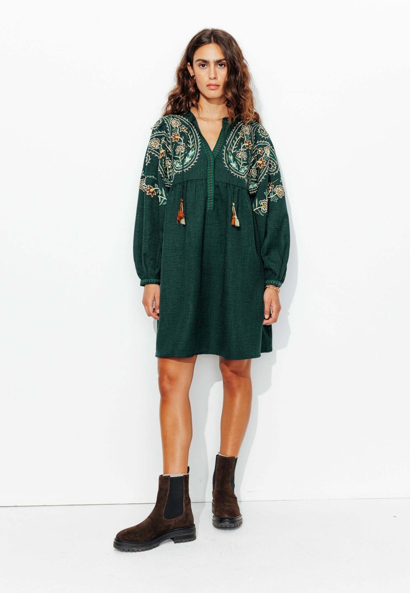 Robe verte brodée avec des manches longues, décolleté en V et des franges à l'avant. Associée à des bottes chelsea marron.