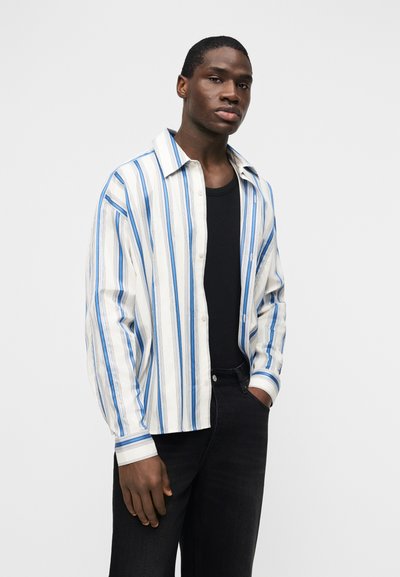 Νεαρός άνδρας που φοράει λευκό και μπλε ριγέ πουκάμισο κουμπωτό πάνω από μαύρο αμάνικο, με μαύρο τζιν, στέκεται με το ένα χέρι στην τσέπη.