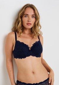 Reggiseno in pizzo blu navy con bordi scallop, supporto con ferretto e spalline regolabili. Presenta motivi floreali e un fiocco in satin al centro.
