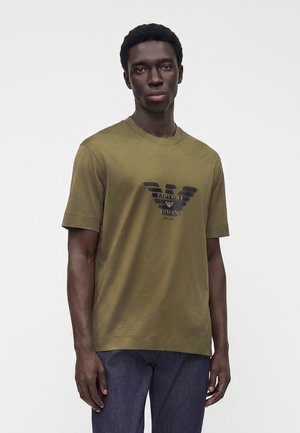 Νέος άνδρας με μια ελιάς πράσινη μπλούζα Emporio Armani και σκούρο τζιν στέκεται μπροστά σε ένα απλό φωτεινό φόντο.