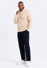 Felpa in pile beige con coulisse, tasca frontale, abbinata a pantaloni cargo scuri e sneakers bianche. Design semplice e funzionale.