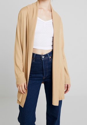 Personne portant un cardigan beige ouvert, un haut court blanc et un jean taille haute bleu foncé sur un fond gris uni.
