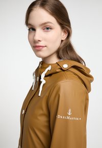 Brun vandtæt jakke med hætte, hvide snore og logo detalje på ærmet. Glat tekstur og moderne design.