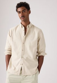 Homme aux cheveux bouclés et moustache portant une chemise beige à boutons et un pantalon clair, debout les mains dans les poches devant un fond uni.