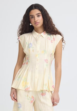 Kvinde med langt krøllet hår iført en ærmeløs, cremefarvet skjorte med knapper og matchende bukser med pastelblomstertryk.