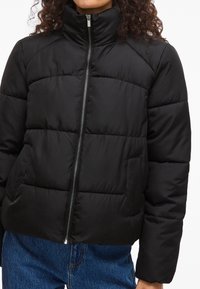 Čierna prešívaná bunda s vysokým golierom, predným zipsom a prešívaným dizajnom. Má bočné vrecká a krátky strih. Materiál pôsobí hladko a leskle.