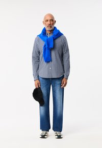 Uomo che indossa un maglione blu sopra una camicia a quadri, jeans di denim larghi, tiene un cappellino nero e indossa scarpe da ginnastica con motivo.