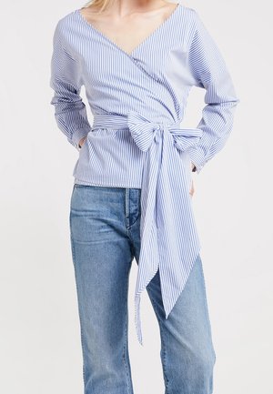 Femme portant un chemisier cache-cœur rayé bleu et blanc avec un grand nœud à la taille et un jean en denim bleu clair, mains dans les poches.