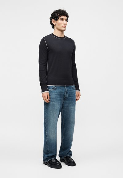 Homme debout portant une chemise noire à manches longues avec des coutures blanches, un jean bleu large et des chaussures noires sur un fond blanc uni.