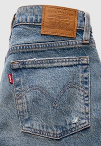 Μπλε τζιν παντελόνι Levi's με πίσω τσέπη που φέρει τη χαρακτηριστική ραφή arcuate, κόκκινη ετικέτα Levi's και ταμπέλα από δέρμα σε ταμπά απόχρωση πάνω από τη μέση.