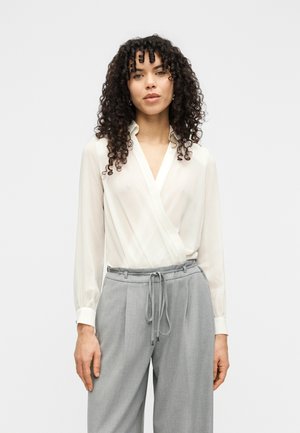 Blusa avvolgente bianca e trasparente con maniche lunghe e colletto appuntito, abbinata a pantaloni a vita alta grigio chiaro con coulisse in vita.