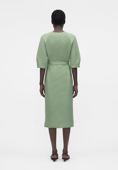 Γυναίκα φοράει μια μίντι μακριά, υφασμάτινη ροφή πράσινου χρώματος φασκόμηλο με μανίκια μέχρι τον αγκώνα και ζώνη, όρθια ξυπόλητη σε λευκό φόντο, πίσω όψη.