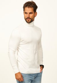 Weißer Rollkragenpullover aus glattem Strickstoff. Figurbetonter Schnitt mit langen Ärmeln und gerippten Bündchen und Saum. Kombiniert mit blauen Jeans.