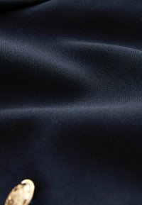 Námořnická tkanina se hladkým, mírně pružným povrchem obsahuje zaoblený, texturovaný zlatý knoflík, který zvýrazňuje spodní pravý roh.