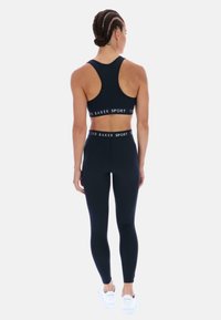 Moteris su pintomis plaukais, vilkinti juodas Ted Baker sportines leggingas ir atitinkamą sportinį liemenėlę, stovinti su nugarą atsukta į priekį ant balto fono.