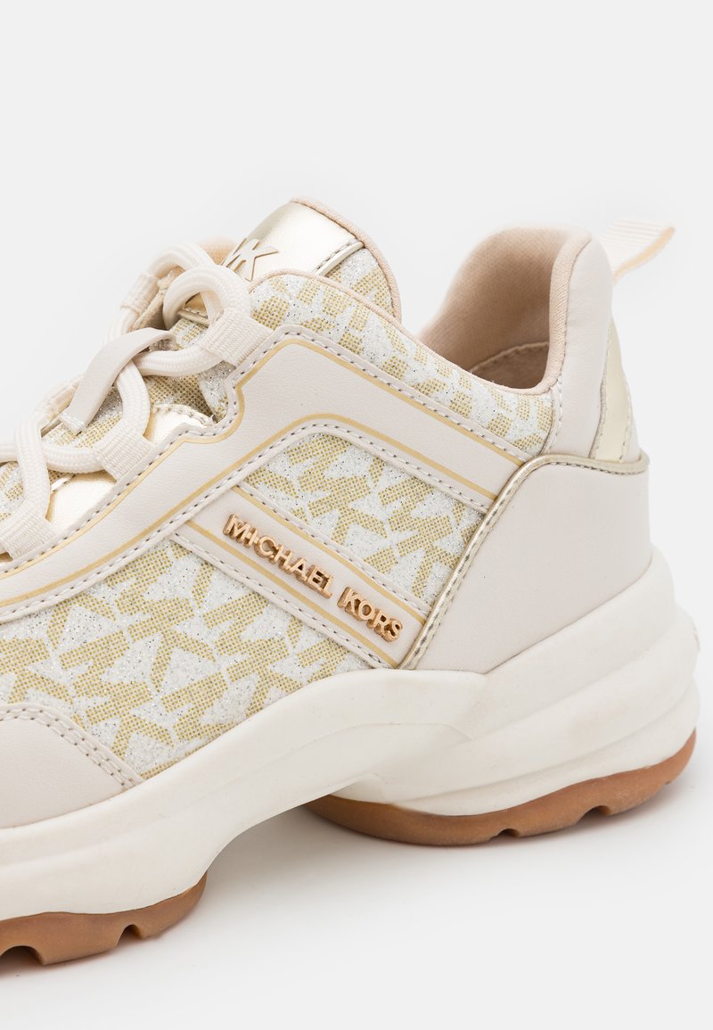 Zapatillas beige con acentos dorados, tejido texturizado con un patrón sutil y una suela blanca gruesa. Logo de la marca en el lateral.
