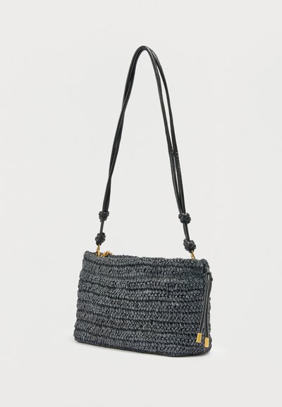 Μαύρη υφαντή τσάντα ώμου με ρυθμιζόμενο λουράκι με κόμπους και χρυσούς φερμουάρ, σε απλό φόντο.