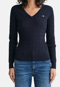 Vrouw in een marineblauwe kabeltrui met V-hals, gedragen met medium-was blauwe jeans, staand met de armen ontspannen langs de zij.