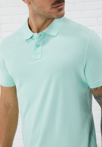 Lys myntgrønn polo skjorte med krage, to knappelukkinger og korte ermer, laget av et teksturert stoff.