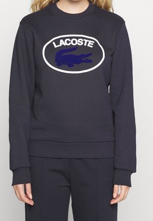 Osoba nosząca ciemnogranatową bluzę Lacoste z białym owalnym logo i granatowym wzorem krokodyla na piersi, w połączeniu z dopasowanymi spodniami.