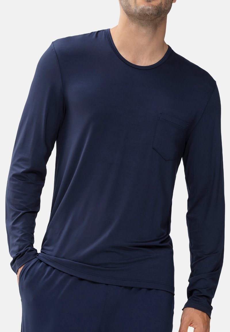 Mężczyzna w ciemnoniebieskiej koszuli z długim rękawem i kieszonką na piersi, z jedną ręką w kieszeni spodni, na jednolitym jasnym tle.