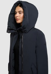 Chaqueta acolchada azul marino con una gran capucha, cremallera frontal y botones a presión, que presenta una superficie texturizada y bolsillos laterales.